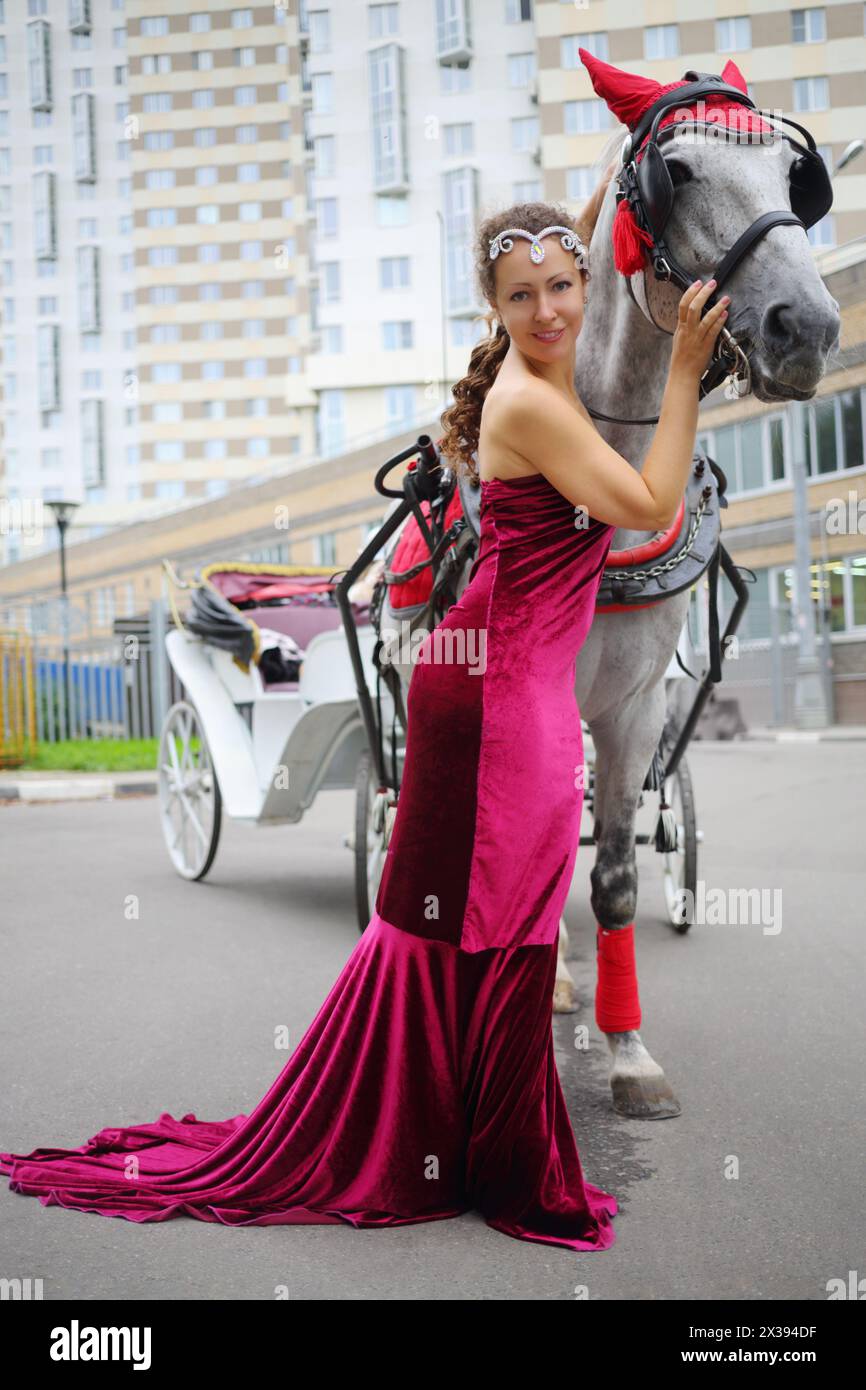 Jolie femme en robe rouge se tient près du cheval avec l'entraîneur près du bâtiment résidentiel Banque D'Images