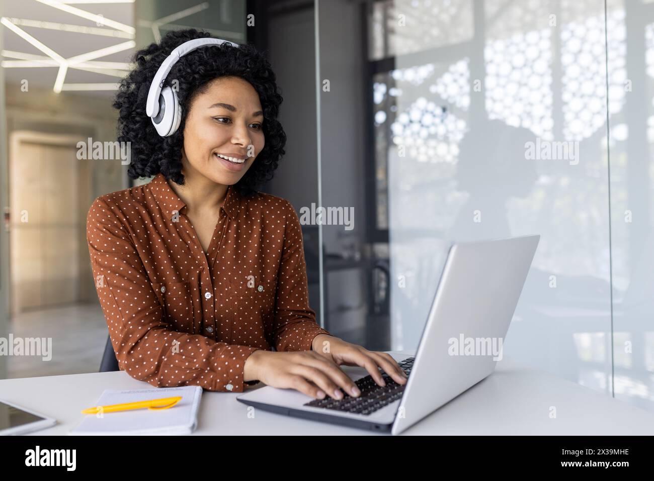 Une jeune femme afro-américaine portant une chemise à pois et des écouteurs sourit alors qu'elle travaille sur son ordinateur portable dans un environnement de bureau moderne. Banque D'Images