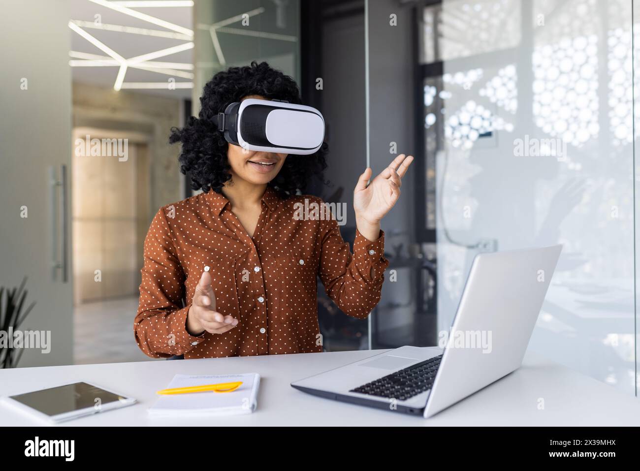 Une jeune femme aux cheveux bouclés s'engage avec la technologie de réalité virtuelle dans un espace de travail moderne. Elle fait des gestes en plein air tout en portant un casque VR, avec un ordinateur portable et un smartphone sur le bureau. Banque D'Images