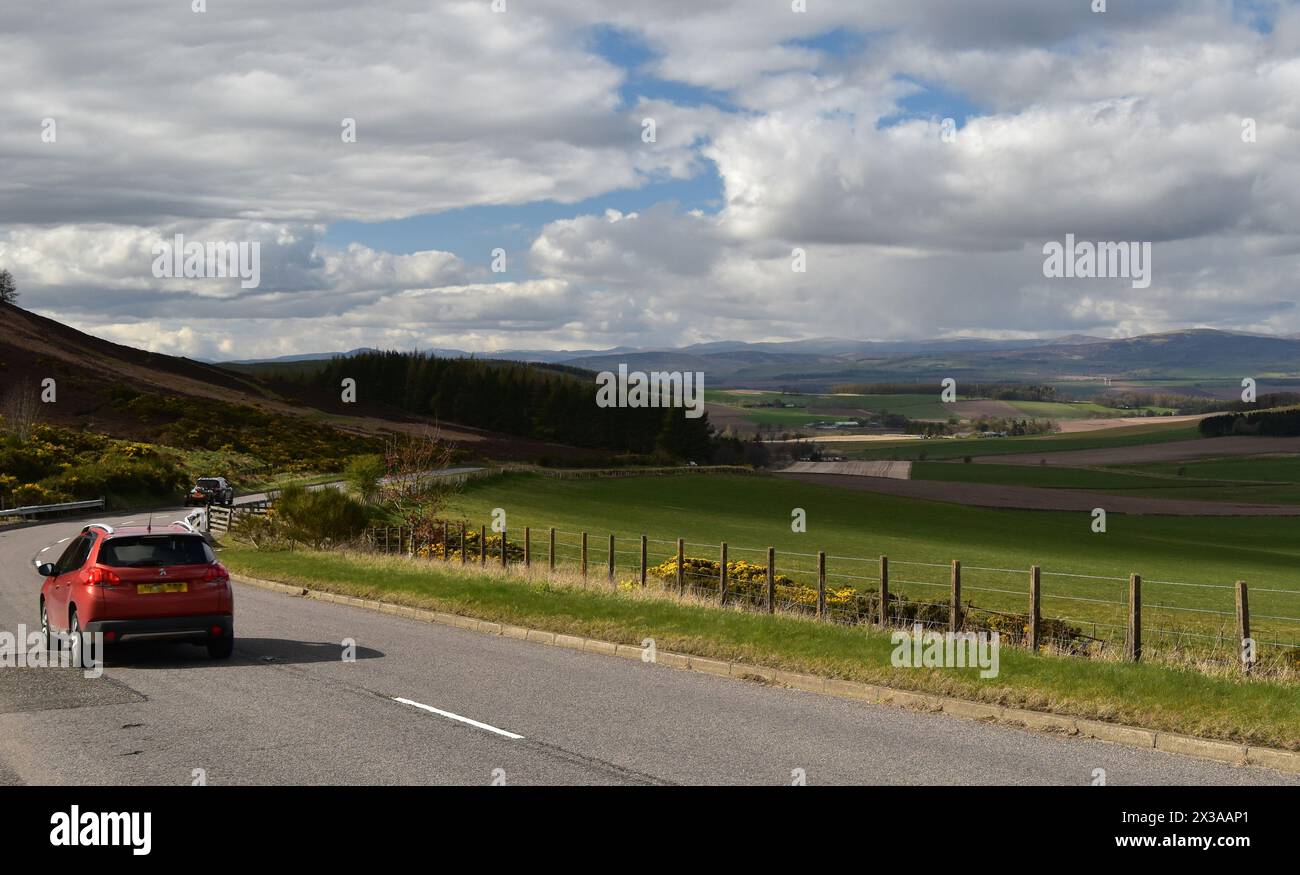 Une voiture rouge conduisant le long de l'A928 en direction de Glamis, devant un point de vue au-dessus de la vallée de Strathmore à Angus, en Écosse, par un jour de printemps brillant. Banque D'Images