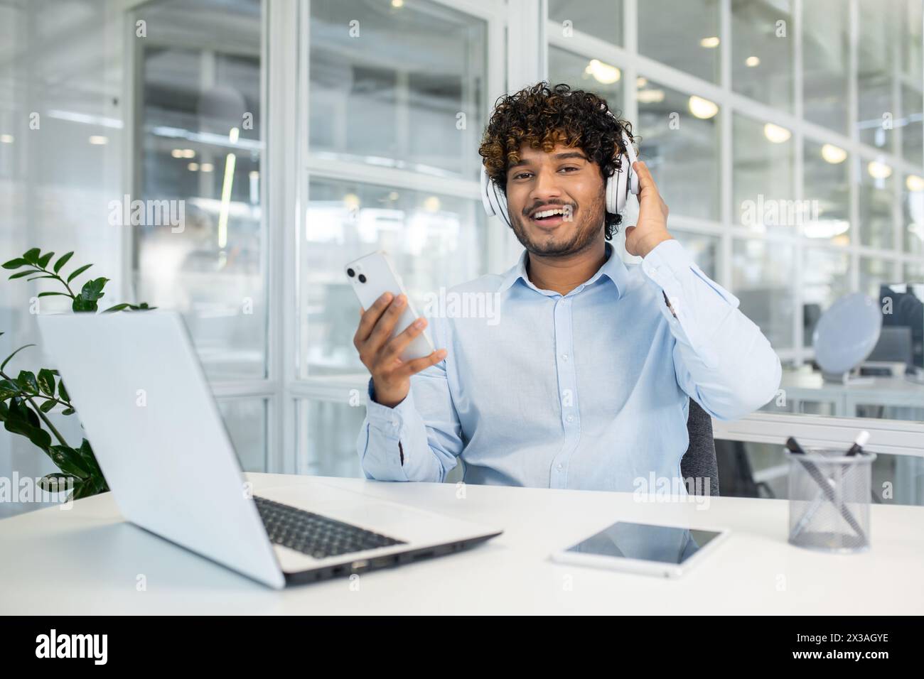 Jeune homme souriant profitant de la musique tout en manipulant des affaires sur son smartphone dans un cadre de bureau lumineux et moderne. Un concept d'équilibre travail-vie privée et de technologie. Banque D'Images