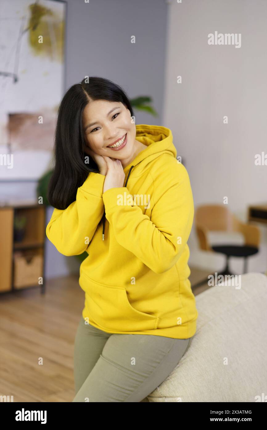 Une femme dans un sweat à capuche jaune sourit et pose pour une photo. La chambre a une atmosphère chaleureuse et accueillante, avec un canapé et une plante en pot à proximité Banque D'Images