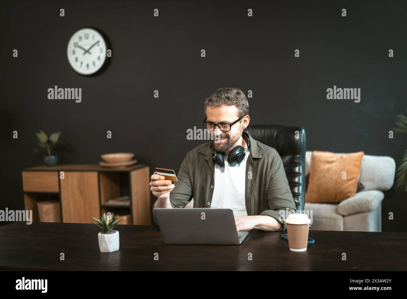 Un homme est assis à un bureau avec un ordinateur portable et une carte de crédit à la main. Il sourit et il s'amuse Banque D'Images