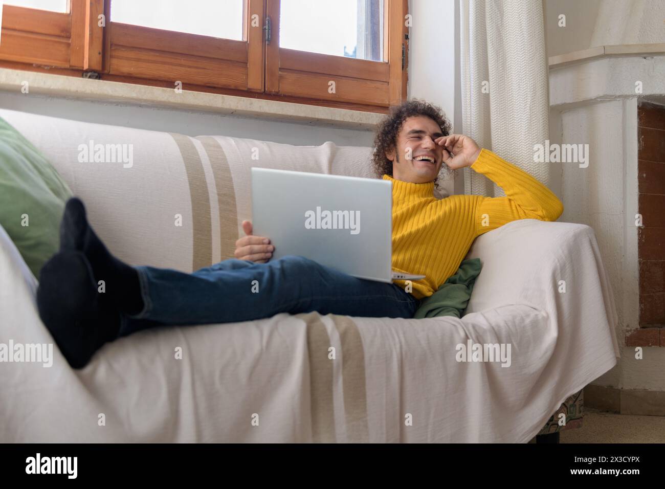 Homme aux cheveux bouclés avec un ordinateur portable dans un pull jaune riant sur le canapé Banque D'Images