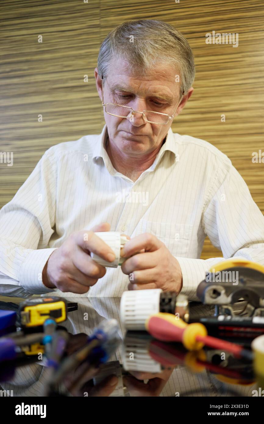 Homme âgé dans des lunettes s'assoit au bureau avec différents outils et détails. Banque D'Images