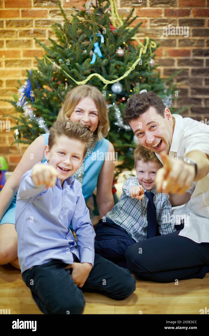 Famille heureuse de quatre est assis sur le sol dans la chambre sous l'arbre de noël, le père et les fils pointent l'index à la caméra, se concentrer sur le visage du père. Banque D'Images