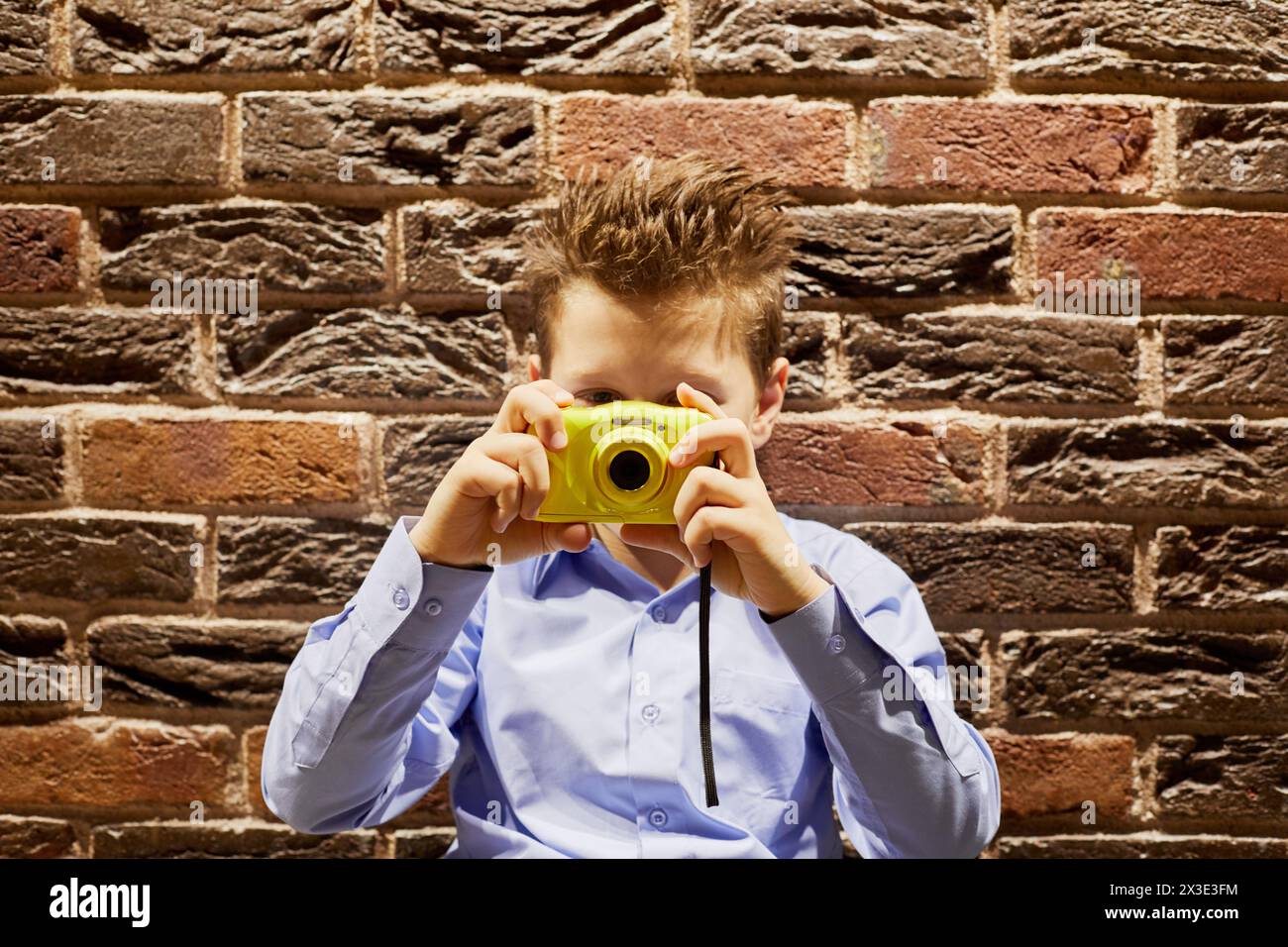 Petit garçon avec caméra jaune contre mur de briques rouges. Banque D'Images