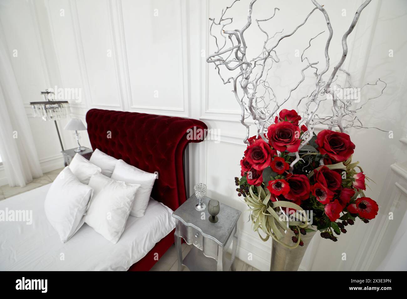 Bouquet de fleurs artificielles, branches, baies dans un vase dans une chambre avec lit rouge. Banque D'Images