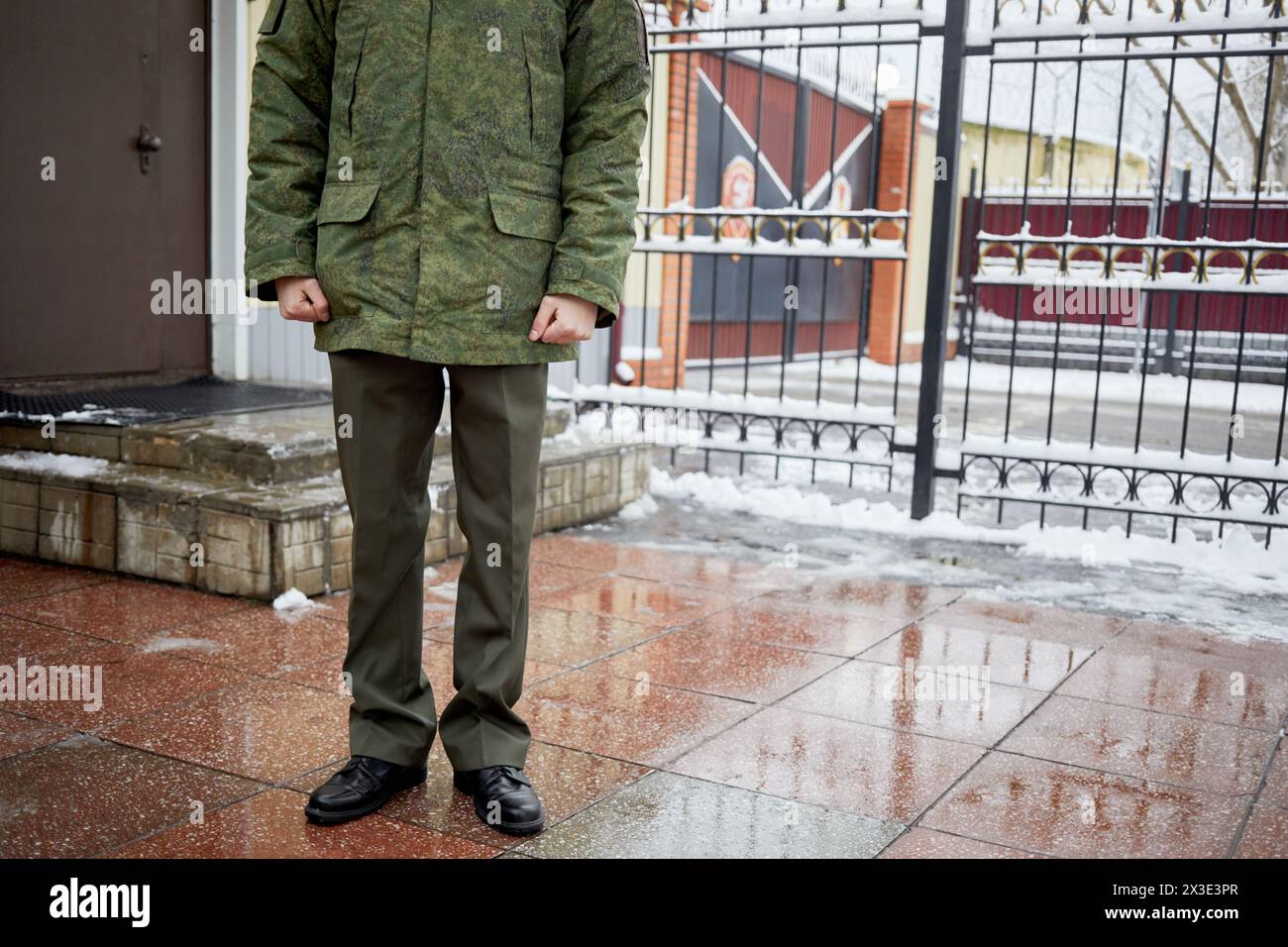 Homme en uniforme militaire se tient près du bureau du poste de contrôle et de la clôture métallique le jour d'hiver. Banque D'Images