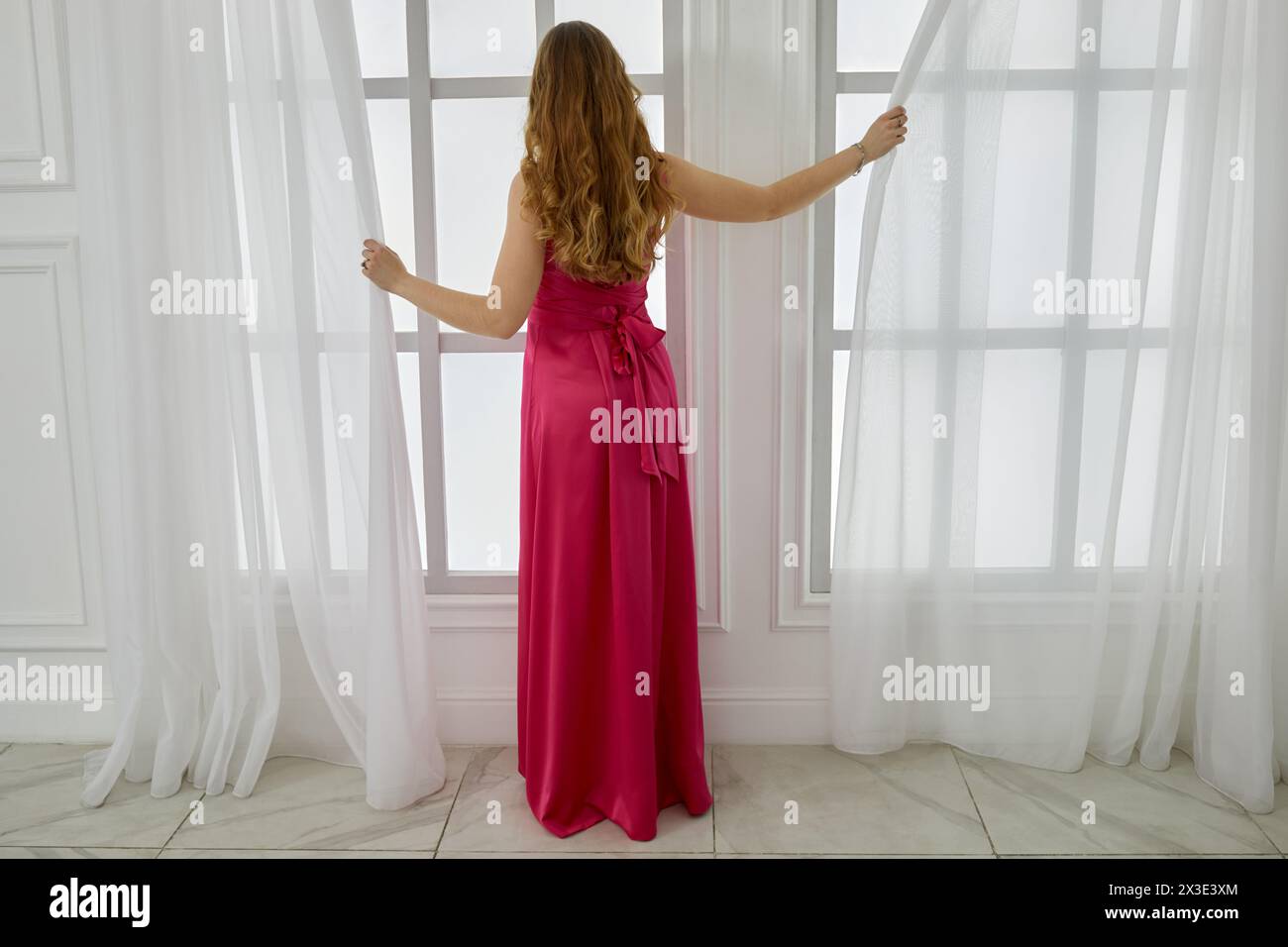 La jeune femme blonde en robe rouge dessine le rideau sur la fenêtre dans la pièce blanche. Banque D'Images