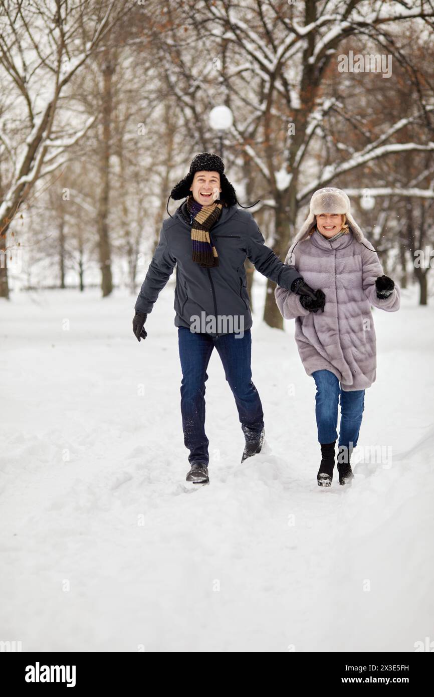 Homme et femme souriants courent en se tenant la main dans un parc enneigé d'hiver. Banque D'Images