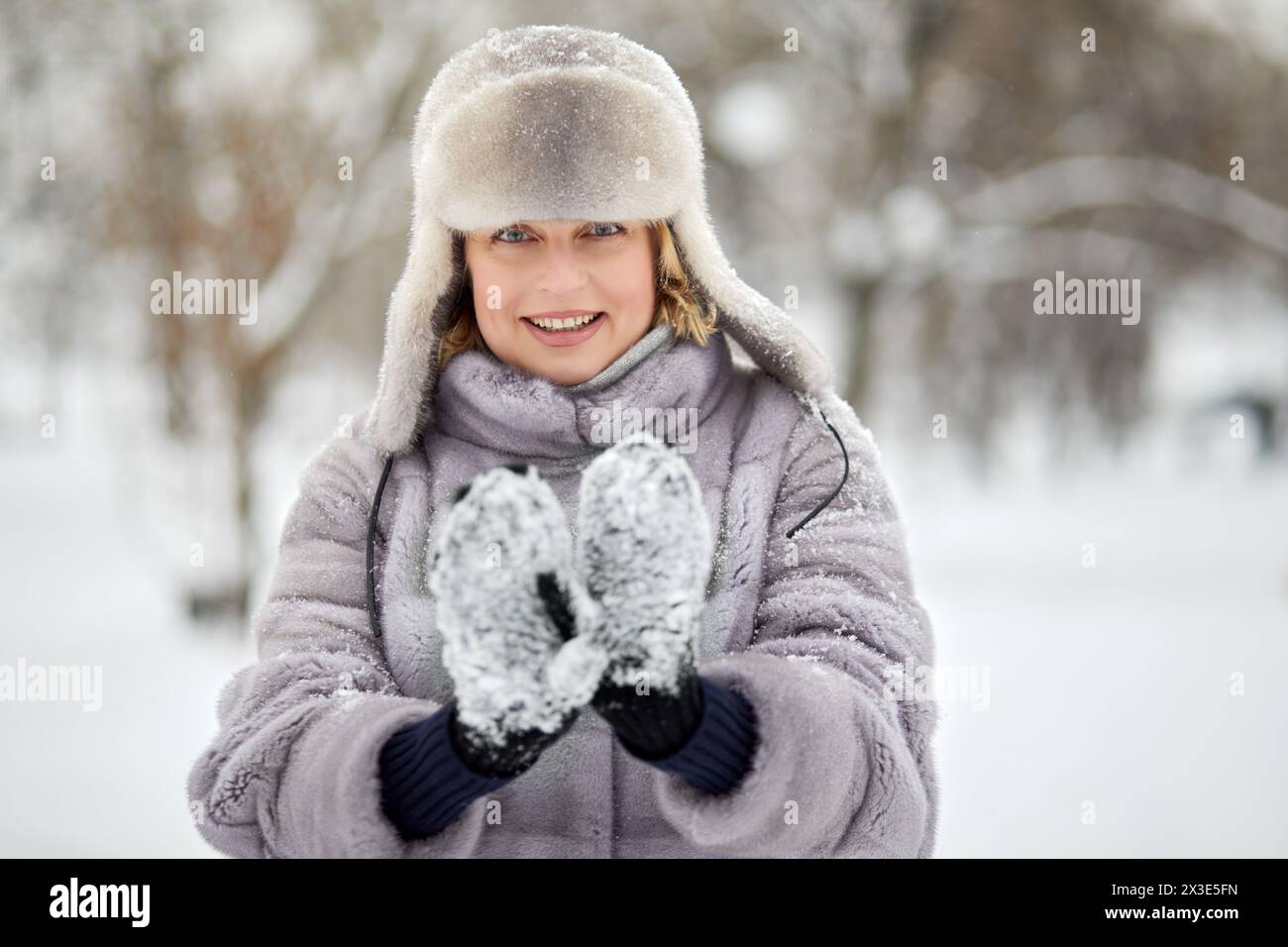 Femme souriante en manteau de fourrure et chapeau montre des mitaines avec de la neige coincée dans le parc d'hiver. Banque D'Images