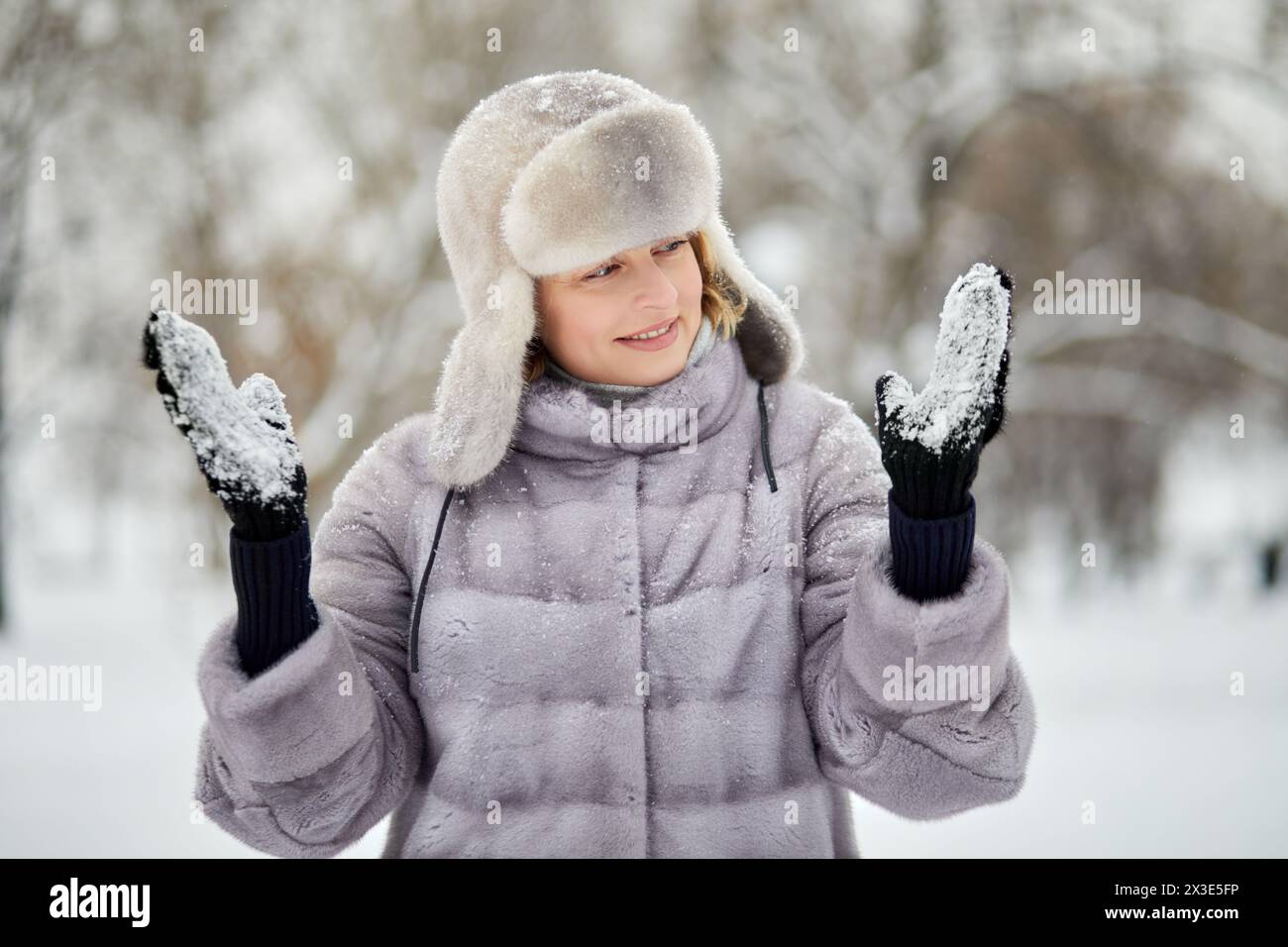 Femme souriante en manteau de fourrure et chapeau se tient regardant des mitaines sur ses mains avec de la neige collée dans le parc d'hiver. Banque D'Images