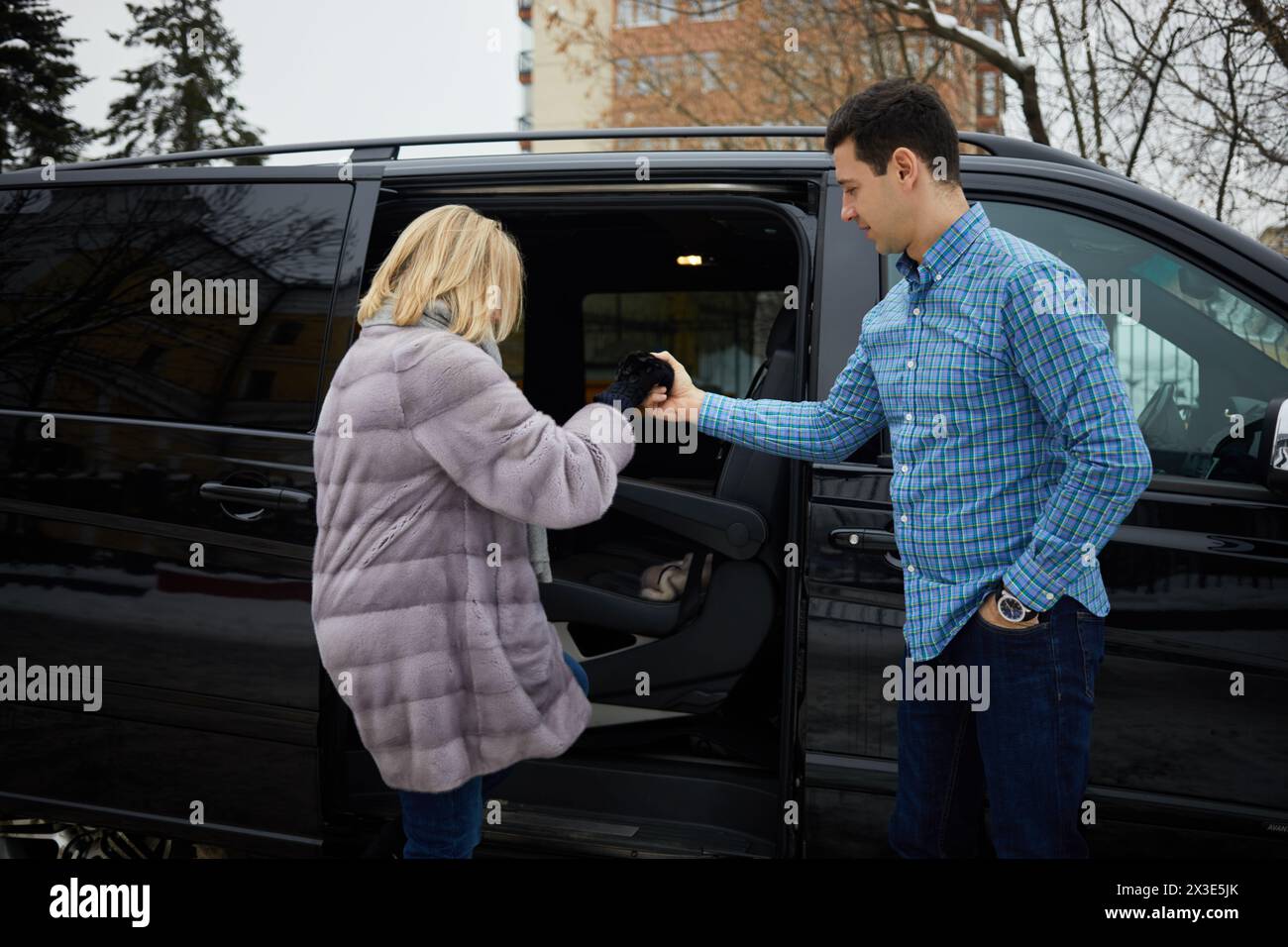 Homme en chemise à carreaux aide la femme en manteau de fourrure pour entrer dans le minivan noir. Banque D'Images