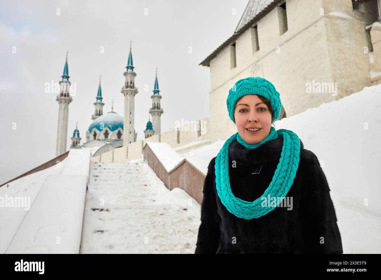 Femme souriante en manteau de fourrure noir se tient au bas des escaliers menant à la mosquée de Kazan Kul Sharif derrière le mur du Kremlin avec la tour le jour d'hiver. Banque D'Images