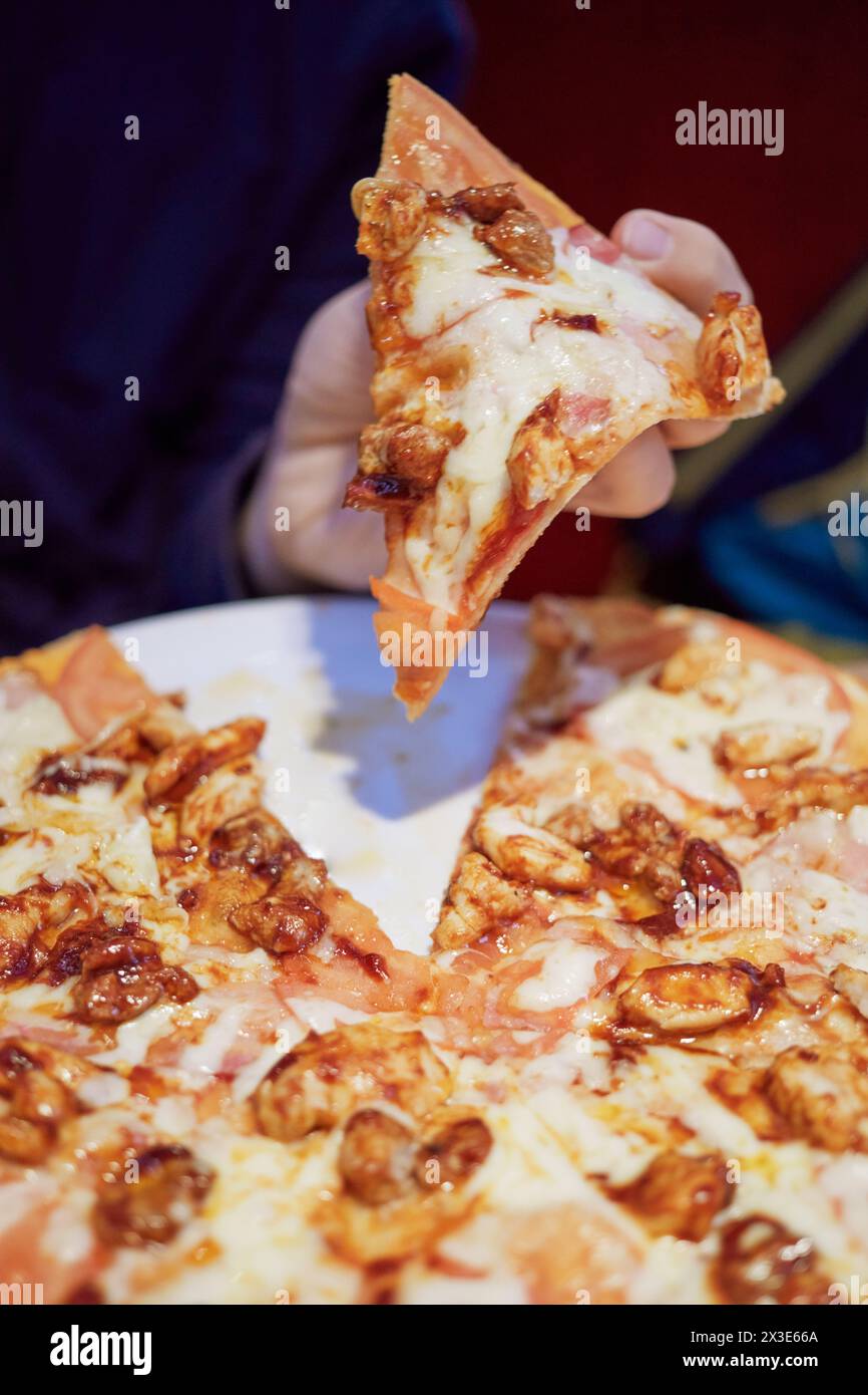 La main humaine tient un morceau de pizza avec jambon, tomate, viande, fromage au-dessus de l'assiette. Banque D'Images