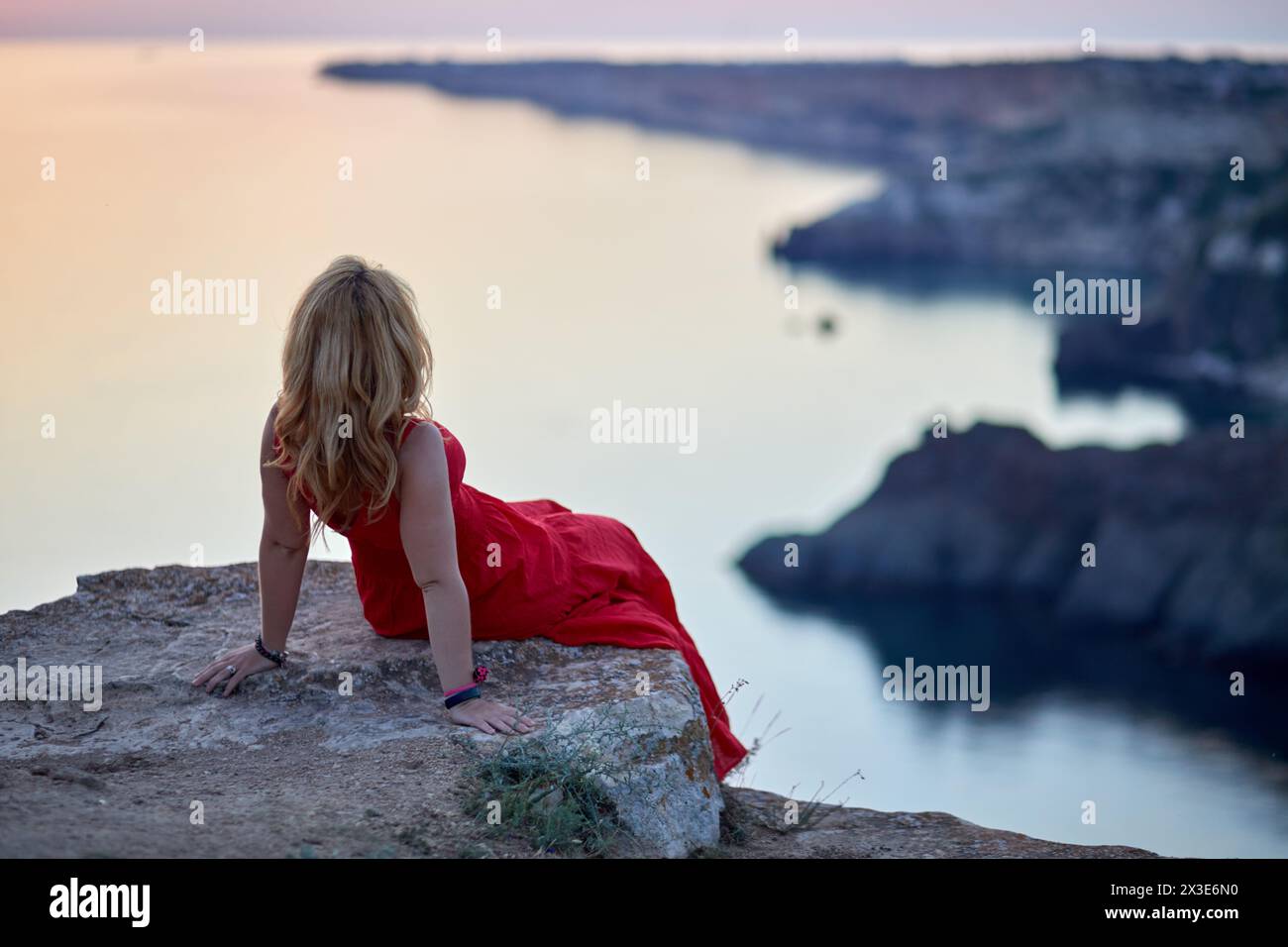 Femme en robe rouge est assis sur le bord de la falaise de pierre penchée en arrière et regardant le bord de mer, vue arrière. Banque D'Images