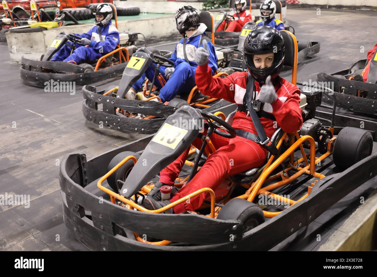 Cinq adultes et un enfant en casques se préparent à la conduite de karting, femme dans les pouces rouges vers le haut, se concentrer sur la femme droite Banque D'Images