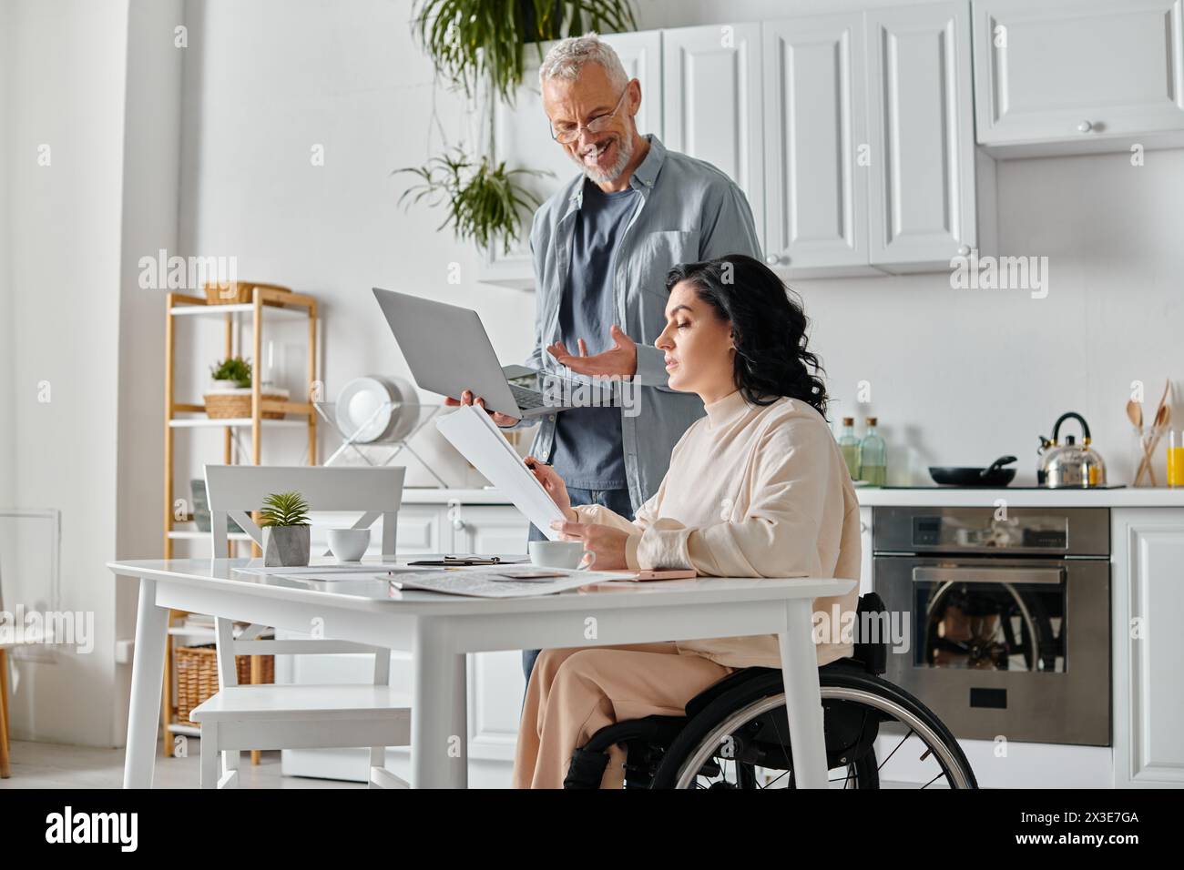 Un homme et une femme dans un fauteuil roulant engrossés dans un écran d'ordinateur portable dans une cuisine confortable à la maison. Banque D'Images