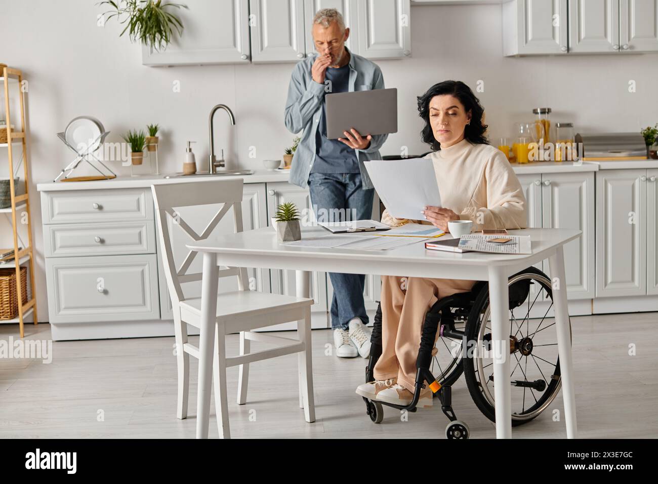 Une femme handicapée en fauteuil roulant interagit avec un ordinateur portable, soutenu par son mari dans une cuisine confortable à la maison. Banque D'Images