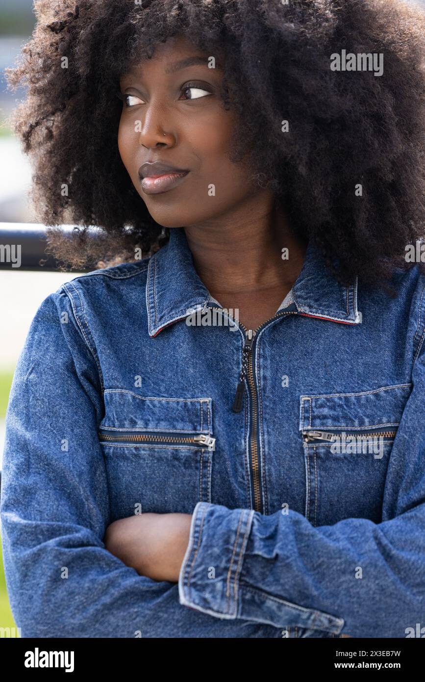 Un gros plan représente une femme noire avec un afro, vêtue d'une veste en denim classique, regardant avec une expression rêveuse. La mise au point douce sur son visage renforce son regard contemplatif, suggérant une pensée profonde ou une aspiration, dans un contexte urbain. Dreamy Black femme en veste denim regardant loin. Photo de haute qualité Banque D'Images