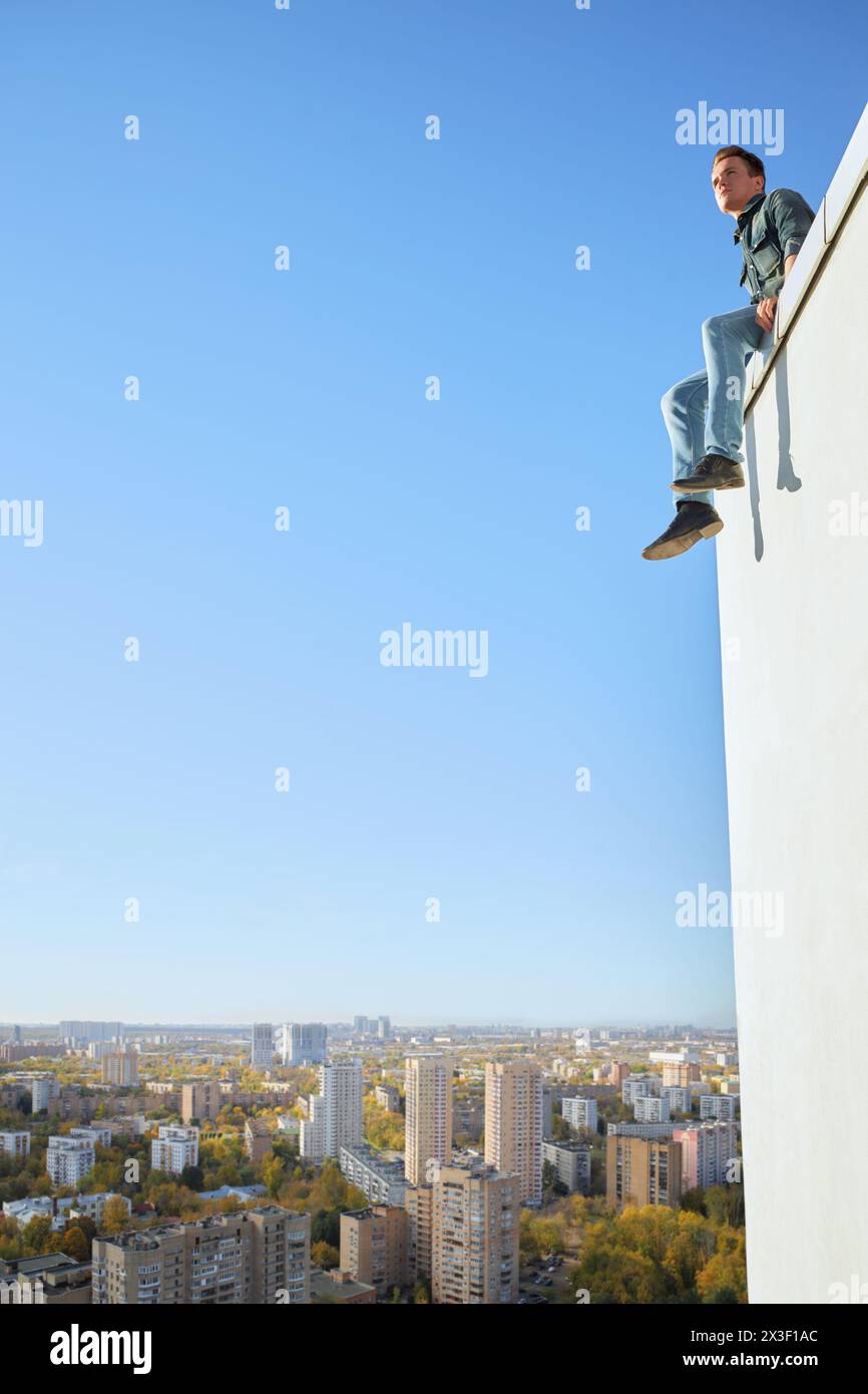 Jeune homme en vêtements denim est assis sur le bord du toit du bâtiment dans la ville ensoleillée Banque D'Images
