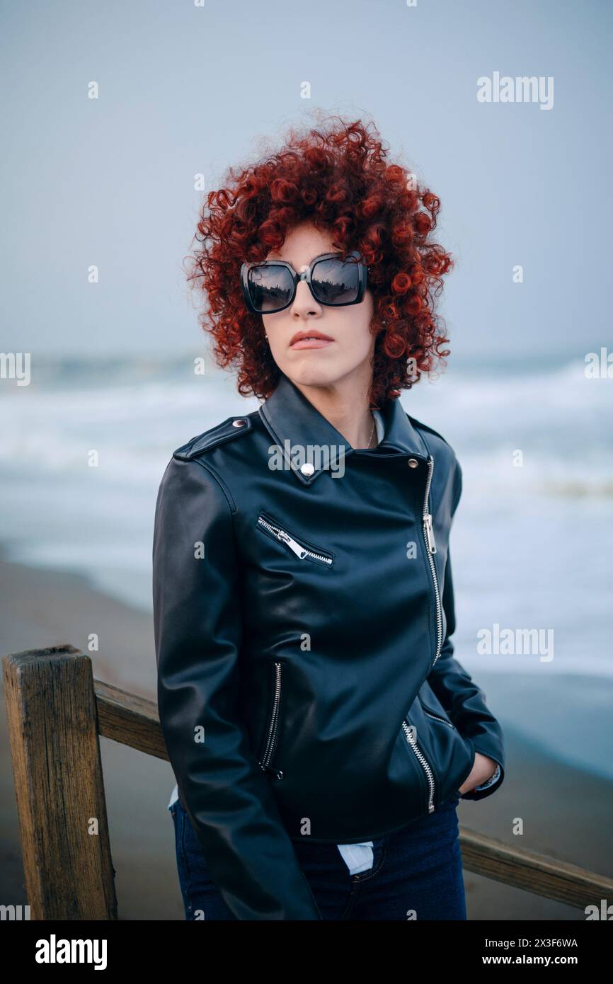 Portrait d'une femme arabe adulte avec les cheveux bouclés rouges dans des vêtements décontractés dans une plage à un jour de pluie Banque D'Images