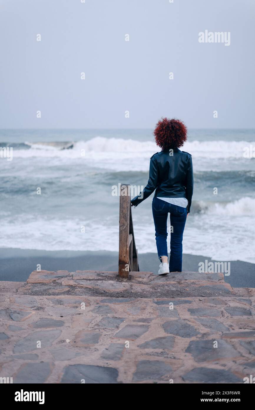 Jeune femme bouclée avec les cheveux roux dans des vêtements décontractés descendre les escaliers à une plage à un jour de pluie Banque D'Images