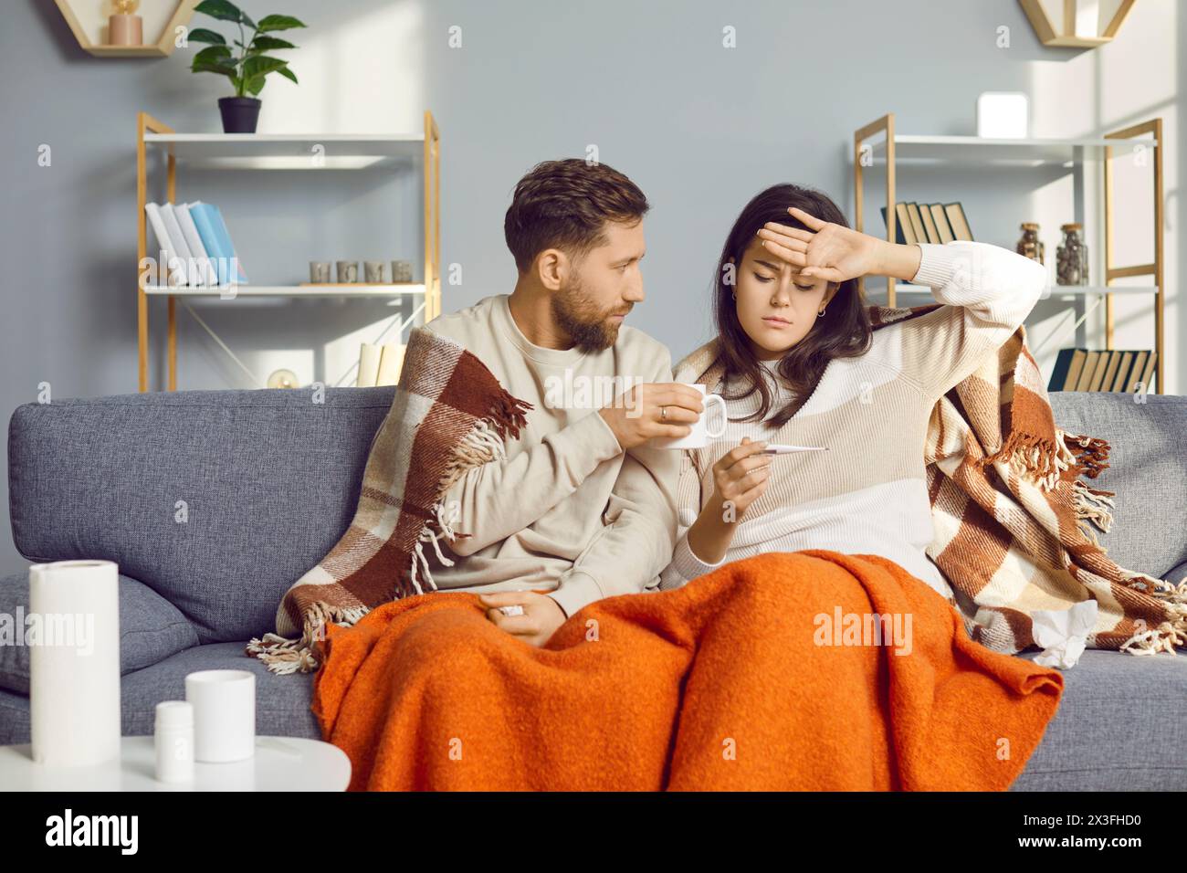 Un homme et une femme malsains sont assis à la maison dans le salon, enveloppés dans une couverture chaude et ressentent les symptômes de la grippe. Jeune couple regarde le thermomètre Banque D'Images