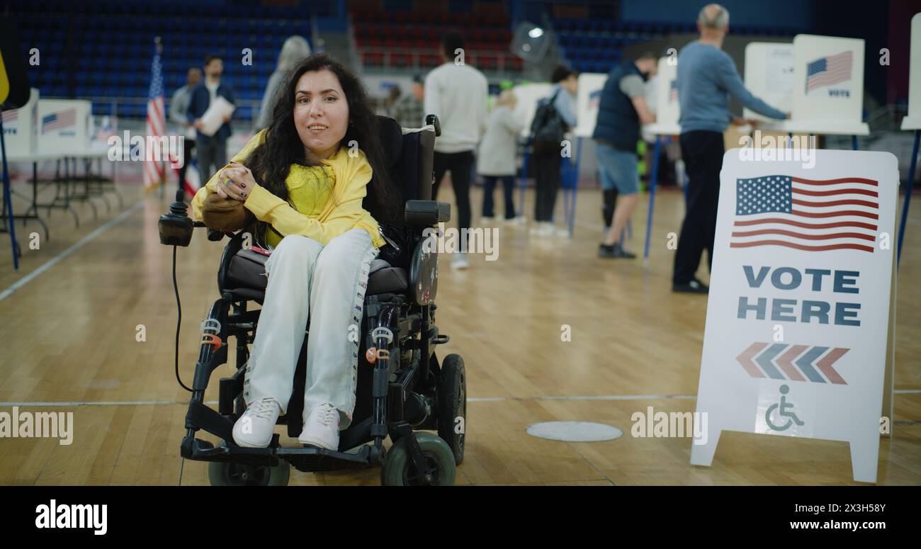 Femme avec SMA en fauteuil roulant électrique sourit et regarde la caméra dans le bureau de vote. Les citoyens américains votent en arrière-plan. Jour des élections nationales aux États-Unis. Concept de devoir civique. Portrait. Banque D'Images