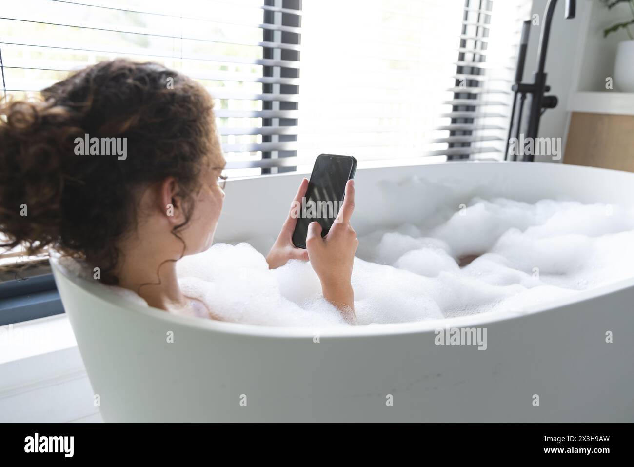 Une jeune femme caucasienne se relaxant dans un bain moussant à la maison, tenant un smartphone, espace de copie Banque D'Images