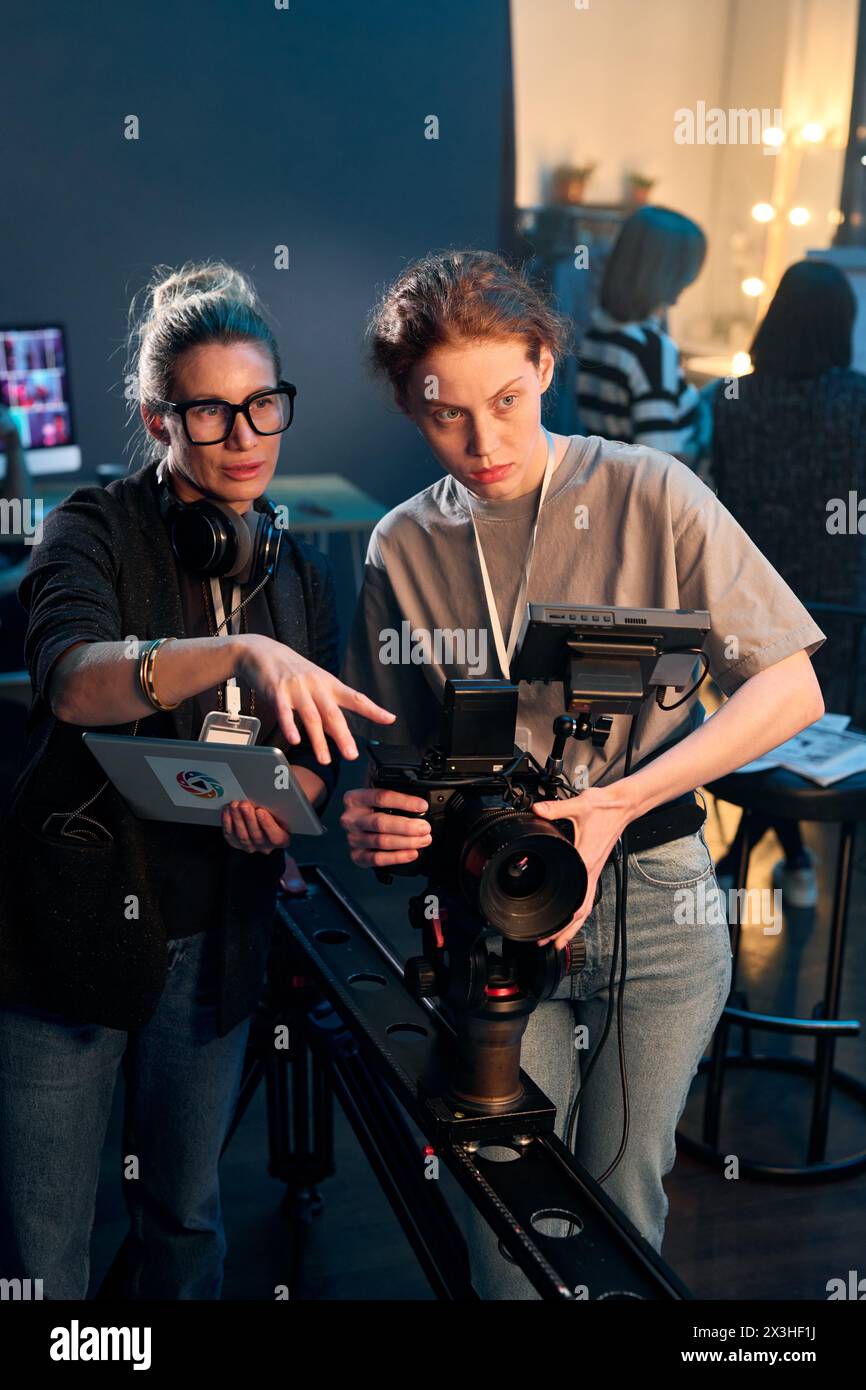 Portrait vertical de camerawoman exploitant la caméra vidéo professionnelle sur la plate-forme avec directrice féminine donnant des instructions Banque D'Images