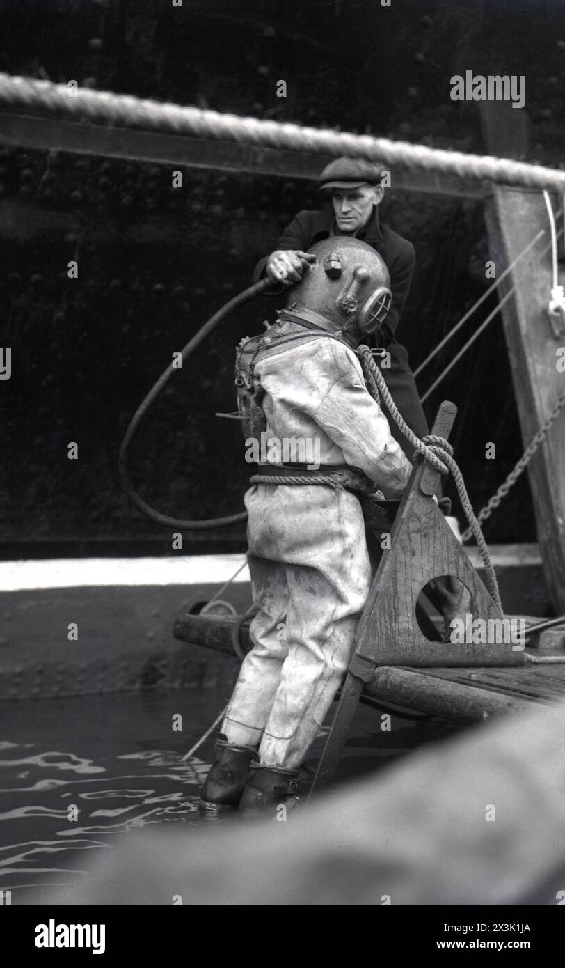 1942, historique, temps de guerre et homme dans le port d'une casquette plate aidant un plongeur en haute mer dans le matériel de plongée de l'époque, sur le point d'entrer dans l'eau, possiblement Southampton docks, Angleterre, Royaume-Uni. Connue sous le nom de robe de plongée standard, de tenue de haute mer ou de tenue lourde, cette combinaison de plongée était utilisée pour des travaux sous-marins profonds et consistait en un casque en métal lourd, en tonneau, en laiton ou en bronze, ajusté à une combinaison imperméable en toile, un tuyau d'air ou respiratoire fixé en surface et des poids en plomb pour contrer la flottabilité. Banque D'Images