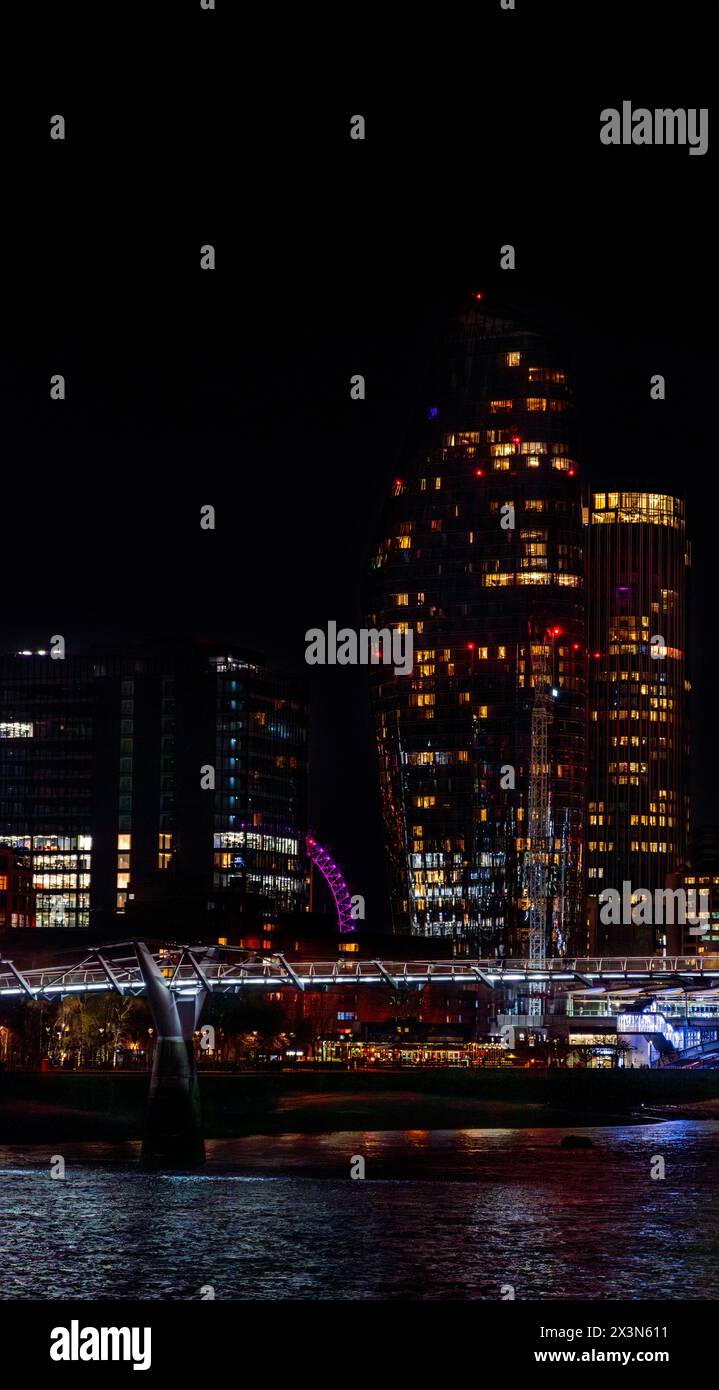 Millennium Bridge illuminé la nuit avec des lumières blanches reflétant ses lumières sur la Tamise et sa rive, avec des gratte-ciel illuminés derrière A. Banque D'Images
