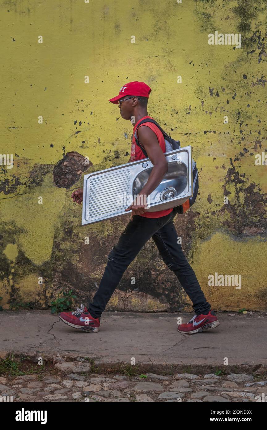 Un jeune homme marchant devant un mur peint en jaune et altéré, portant un évier de cuisine dans les ruelles de la vieille ville, Trinidad, Cuba. Banque D'Images
