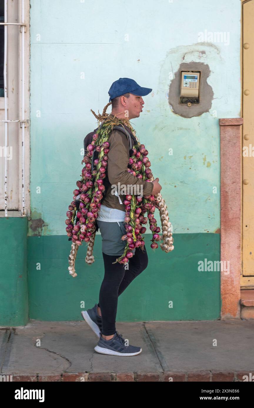 Un vendeur de rue marchant devant un mur altéré, transportant des oignons et de l'ail dans les ruelles de la vieille ville, Trinidad, Cuba. Banque D'Images