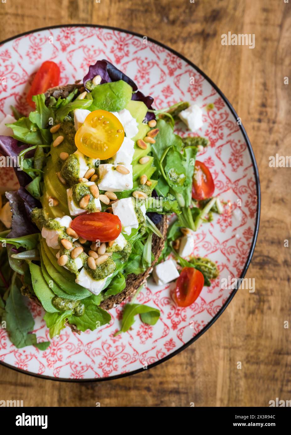 Gros plan vue de dessus de l'avocat avec fromage feta, tomates, pignons de pin, pesto et salade sur pain brun sur une assiette sur une table en bois Banque D'Images