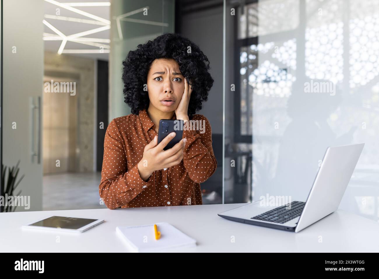 Femme d'affaires afro-américaine dans un bureau moderne, l'air choquée en lisant un message sur son smartphone. Ordinateur portable et documents sur son bureau. Banque D'Images