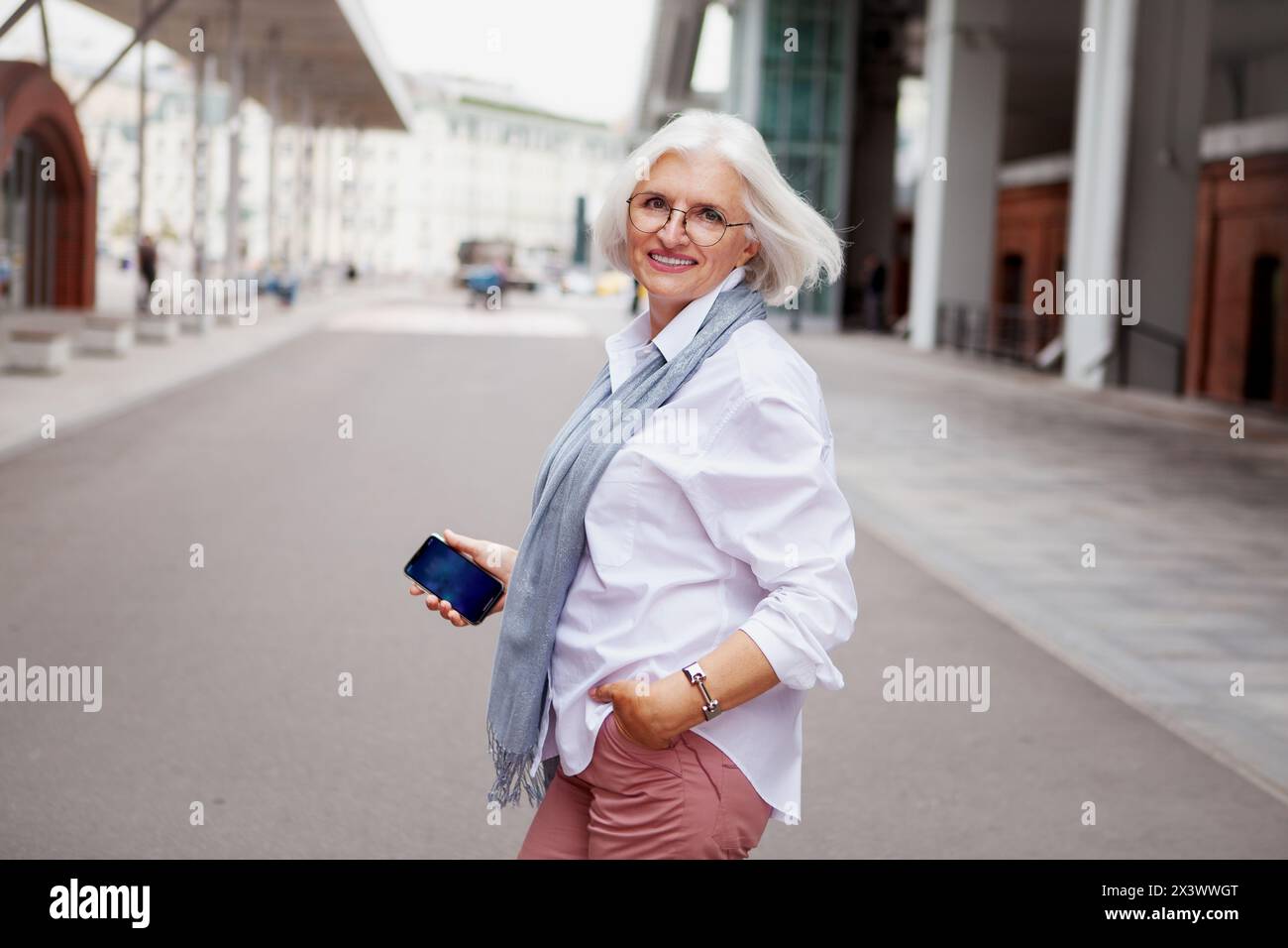 Portrait femme senior souriante heureuse avec les cheveux gris dans la rue de la ville portant la chemise blanche de tenue à la mode exsudant le style et la confiance. Banque D'Images