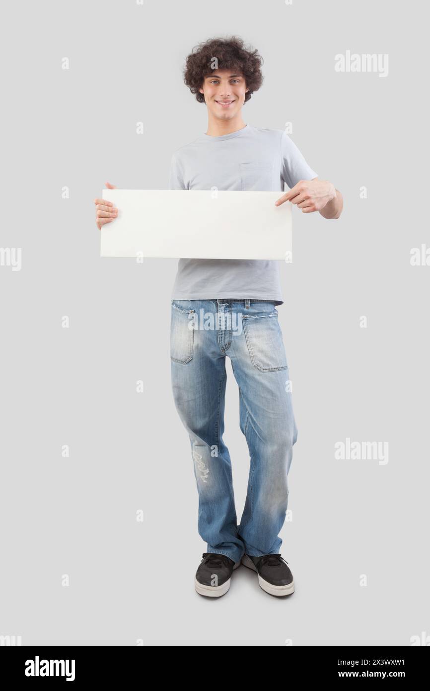 Jeune homme, souriant et beau, pointant avec le doigt pour montrer un panneau blanc vierge, isolé sur fond gris. Espace de copie de l'écriteau pour le texte ou le journal Banque D'Images