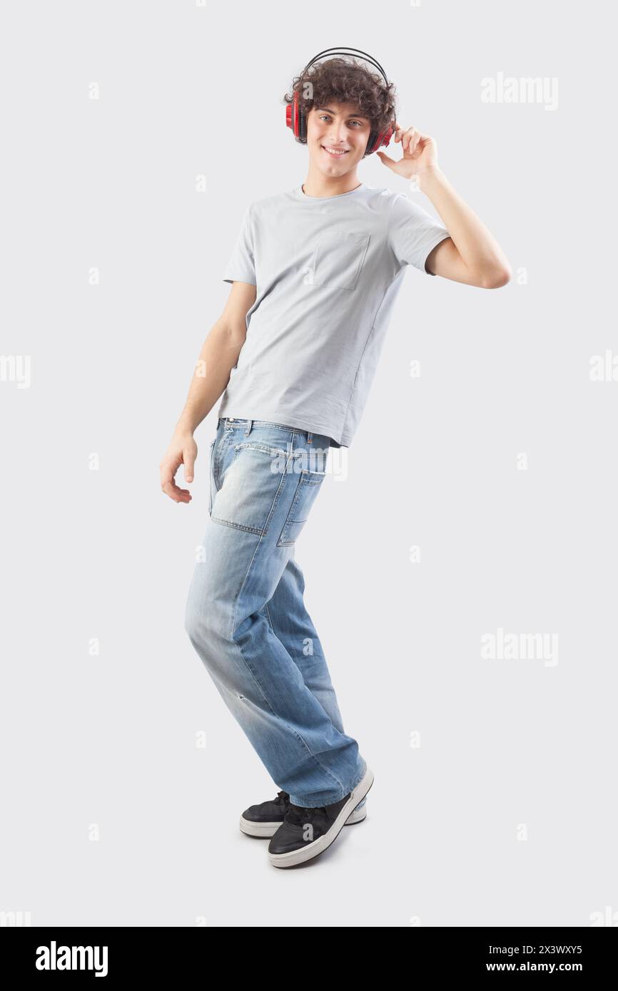 Jeune homme beau et souriant portant des écouteurs pour écouter de la musique et danser tout en regardant dans l'appareil photo, corps entier isolé contre une grotte grise Banque D'Images