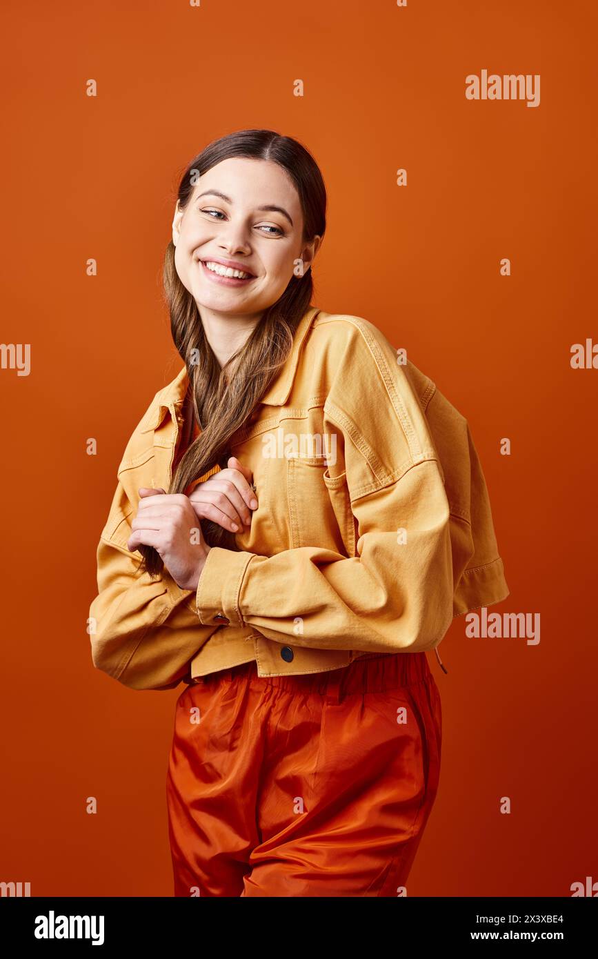 Une jeune femme élégante dans la vingtaine, debout avec confiance, les bras croisés et un sourire éclatant, sur fond de studio orange. Banque D'Images