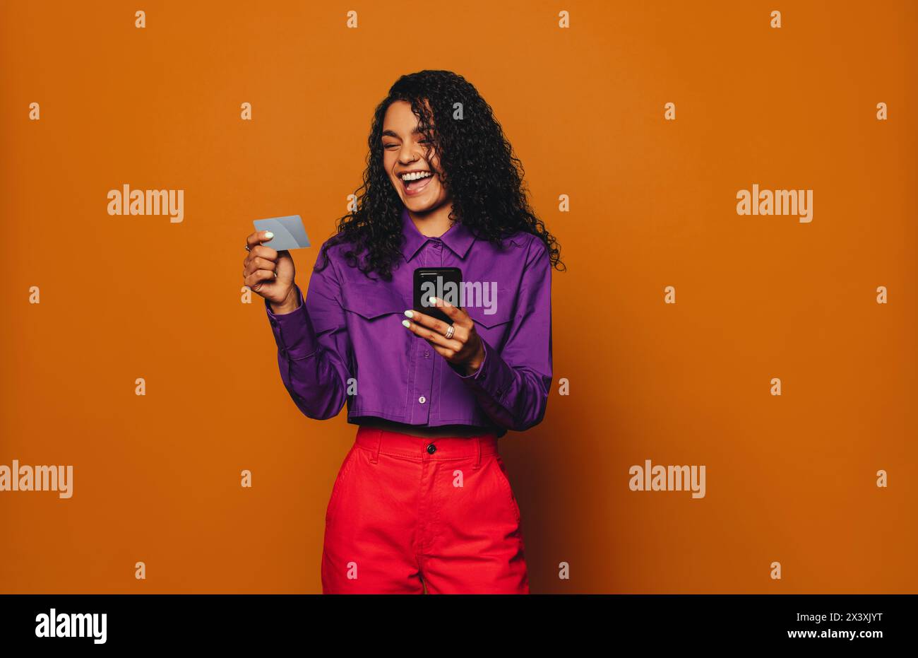 Femme heureuse dans des vêtements décontractés sourires, debout sur un fond orange. En utilisant son smartphone pour faire des achats en ligne, elle paie avec une carte de crédit. Mo Banque D'Images
