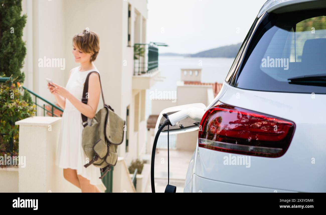 Femme voyageuse avec un smartphone et un sac à côté de la voiture électrique de charge Banque D'Images