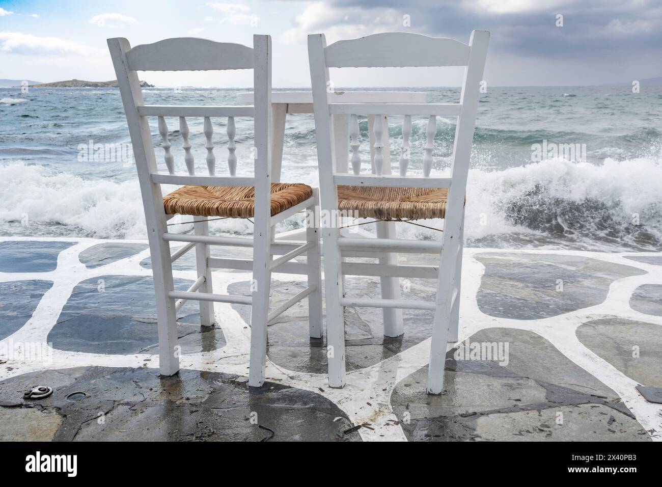 Deux chaises grecques traditionnelles, peintes en blanc, sur un patio en pierre au bord de l'eau accueillant le surf et la vue sur la mer Égée sur l'île... Banque D'Images