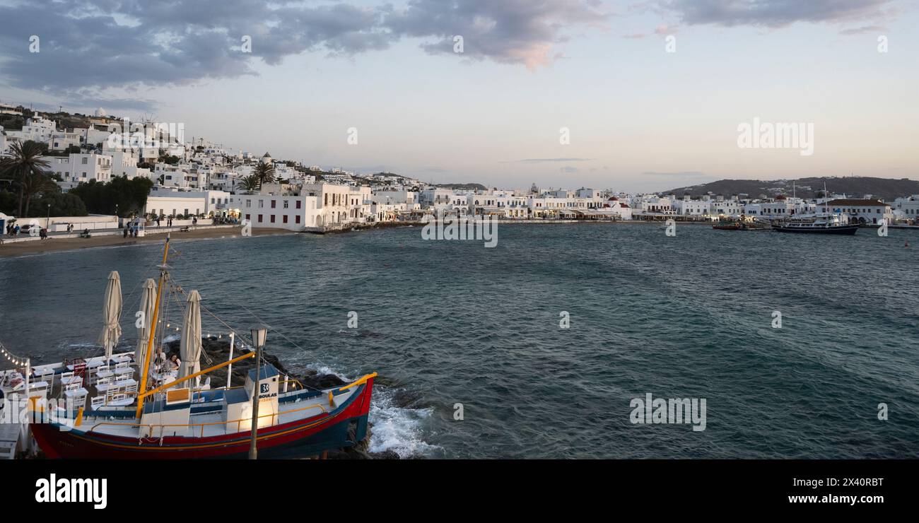 Restaurant en bord de mer dans un bateau le long du rivage et une vue sur les bâtiments blanchis à la chaux dans le paysage urbain de Mykonos au crépuscule Banque D'Images