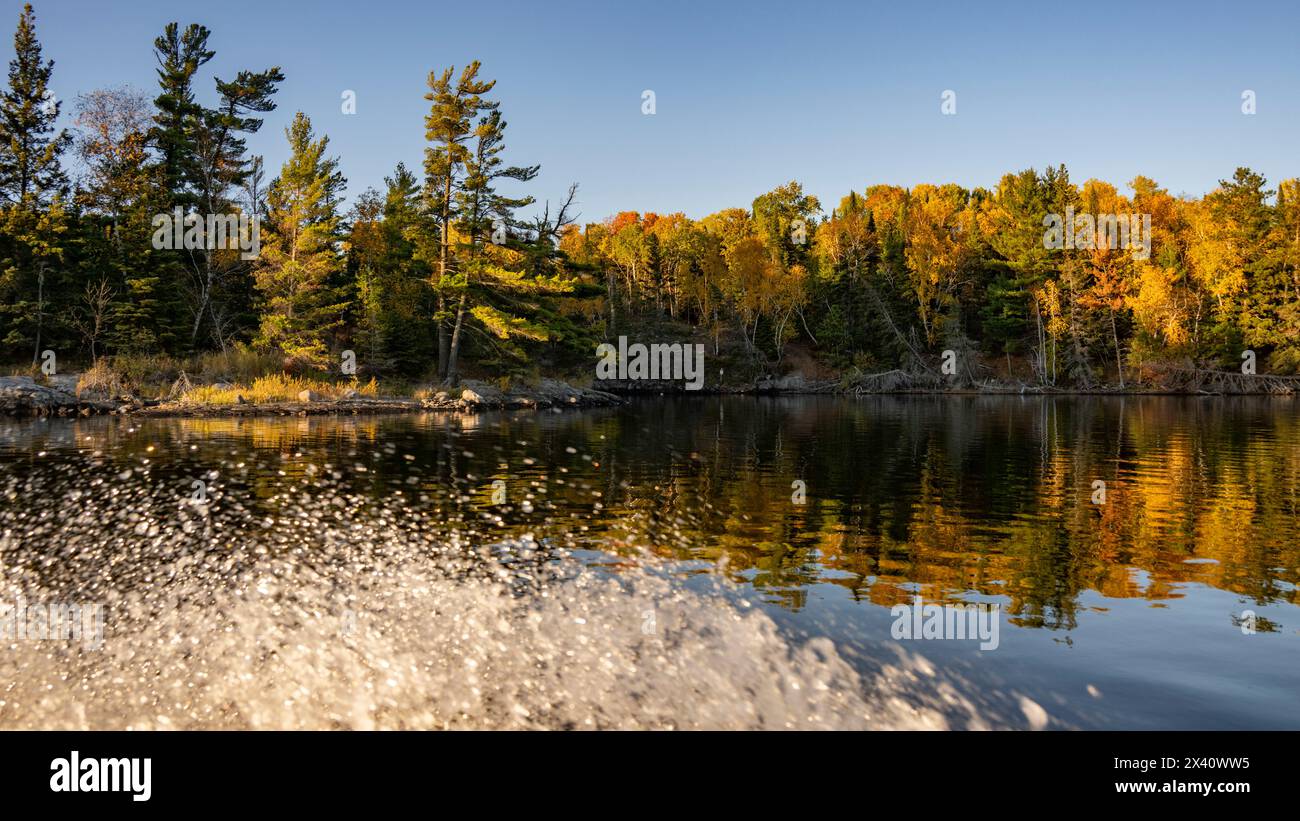 Les éclaboussures d'eau d'un bateau sillonnant un lac tranquille avec un feuillage d'arbres colorés en automne se reflètent dans une image miroir le long du rivage Banque D'Images