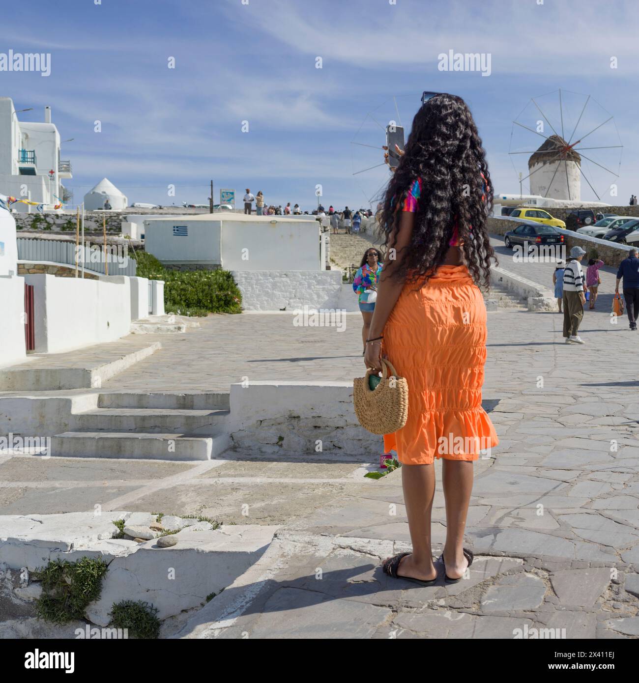 Les touristes apprécient le front de mer et les moulins à vent emblématiques historiques de Mykonos, avec une femme photographiant une scène avec son smartphone au premier plan Banque D'Images