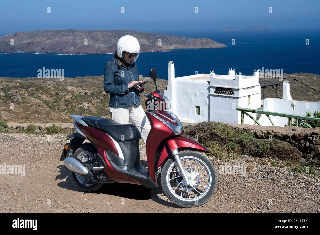 Femme en voyage à moto sur la route vérifie son téléphone intelligent avec une vue sur la côte et la mer Égée ; Ano Mera, sud de la mer Égée, Grèce Banque D'Images