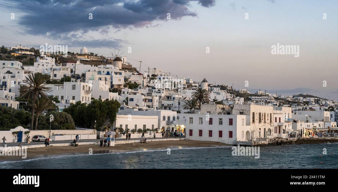 Les gens apprécient le front de mer au crépuscule avec des bâtiments blanchis à la chaux et des palmiers sur une colline sur l'île de Mykonos Banque D'Images